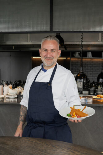 David Legrand, an executive chef, stands in a modern kitchen wearing a white chef's coat and dark apron, holding a plated gourmet dish and smiling confidently.