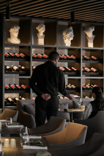 A waiter in a dark uniform stands in front of an elegant wine wall display, featuring shelves lined with bottles and classical sculptures, in a stylish restaurant.