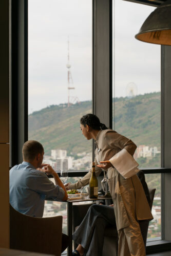 guests enjoy fine dining at Dumas restaurant, located on the 36th floor of Pullman Tbilisi Axis Towers. The window offers an impressive panoramic view of Tbilisi’s landmarks, with the iconic TV tower on Mtatsminda and the cityscape in the background—a perfect spot for an elevated culinary experience.რესტორნის მიმტანი ღვინოს აწვდის სტუმარს დიუმას რესტორანში, პულმან თბილისში, 36-ე სართულზე. ფანჯრიდან ჩანს თბილისის პანორამული ხედი, მთაწმინდის სატელევიზიო ანძით და ქალაქის ხედით